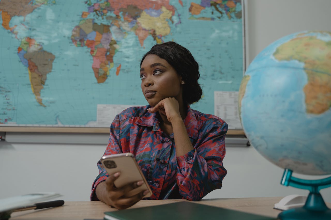 heros-img A teacher in a classroom, pondering with a smartphone. Educational setting with a globe and map.