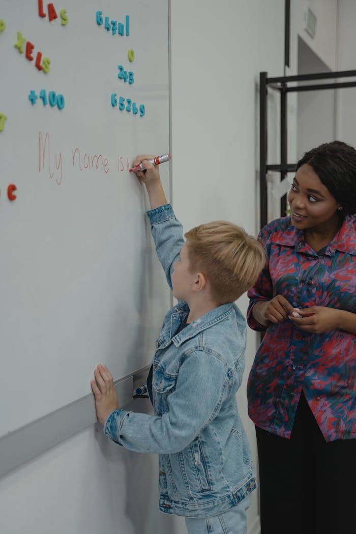 A teacher helps a young student write on a whiteboard during a classroom lesson.